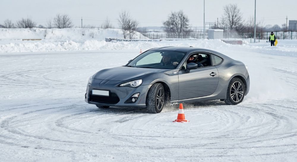 Сіре спортивне авто дрифтує на сніговому автодромі навколо конуса.
